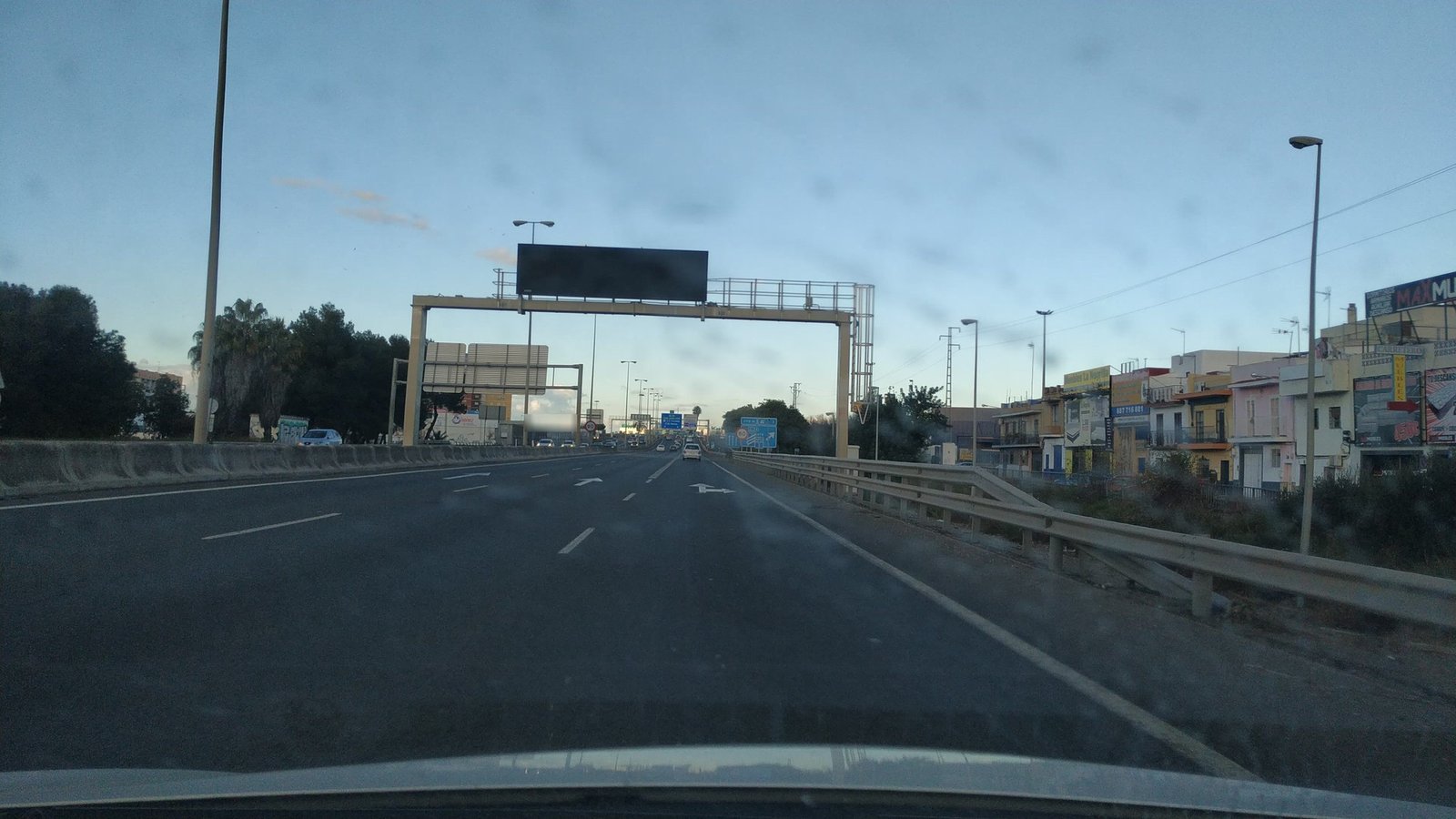 Autovía de varios carriles al acercarse a una ciudad, con un pórtico sobre la calzada y un panel rectangular oscuro, apagado